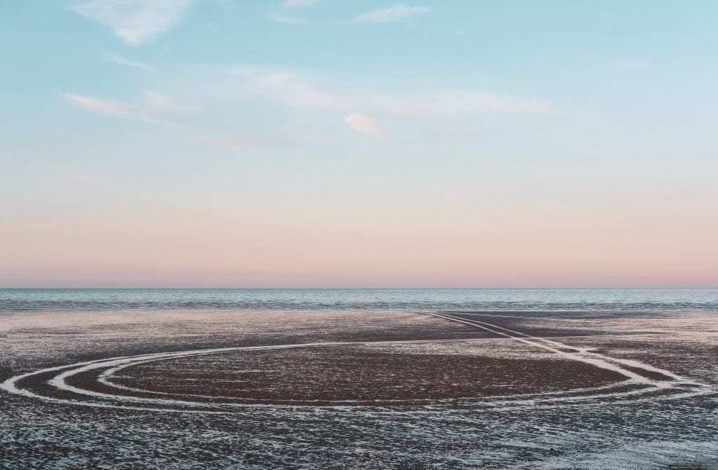 Tyre tracks leading into the sea at Dungeness beach during a pastel sunset by Rachel Groom
