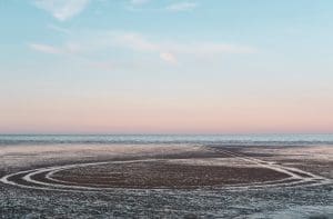Tyre tracks leading into the sea at Dungeness beach during a pastel sunset by Rachel Groom