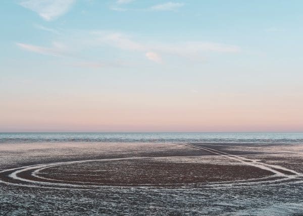 Tyre tracks leading into the sea at Dungeness beach during a pastel sunset by Rachel Groom