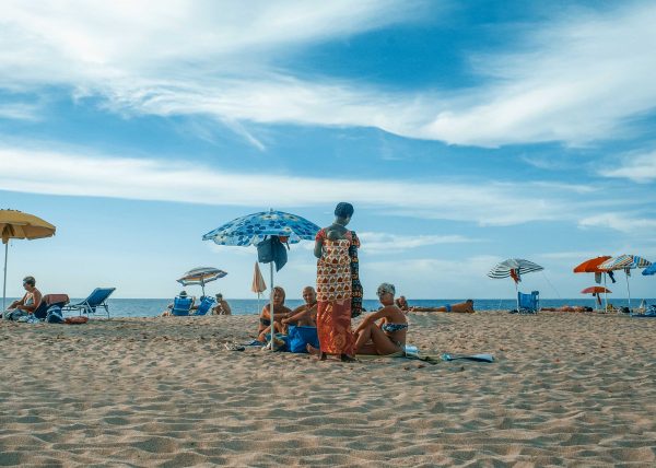 Colourful beach umbrellas and sunbathers on the sandy shore of Sardinia, Italy, under a blue summer sky. Travel photography by Rachel Groom.