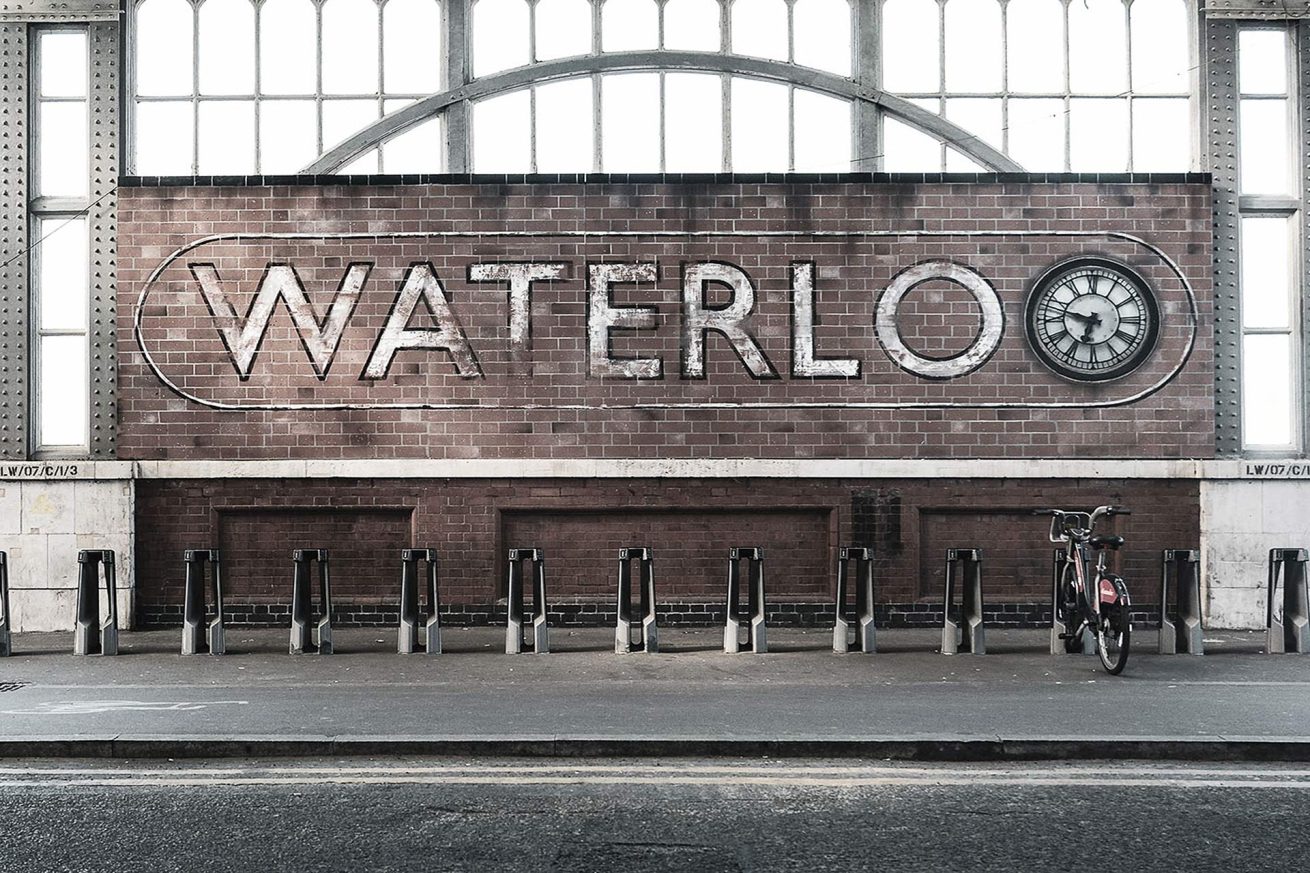 Bike rack in front of a vintage brick wall sign reading 'Waterloo' at Waterloo Station in London, with an old clock on the wall. Photo by Rachel Groom.