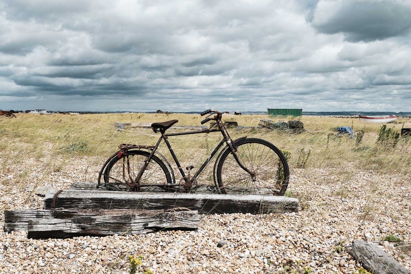 Rusty vintage bicycle leaning on weathered timber in a pebbled field at Dungeness, Kent – photo by Rachel Groom Photography